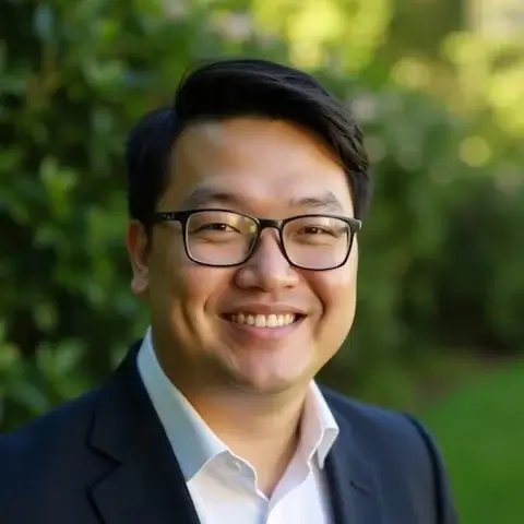 一位男性的专业肖像，戴着眼镜，穿着深色西装和白衬衫，在户外绿植背景前微笑。